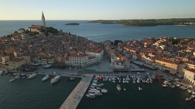 High-angle aerial drone shot capturing Rovinj&rsquo;s lively harbor, moored boats, central waterfront square, and historic peninsula crowned by the Church of St Euphemia bell tower in warm golden hour light