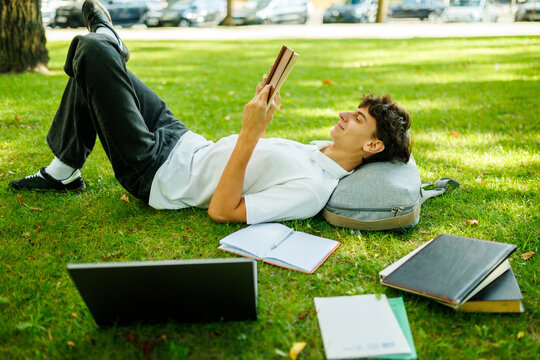 Smiling caucasian student reading a book while lying on the park grass.