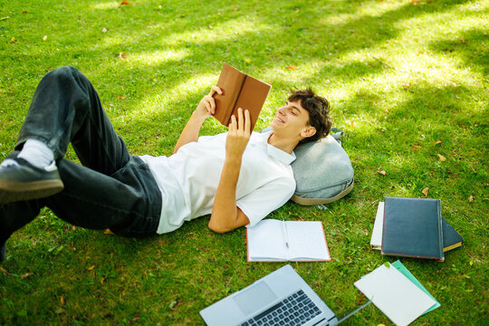 Happy teenage boy reading an educational book on the green outdoor grass.