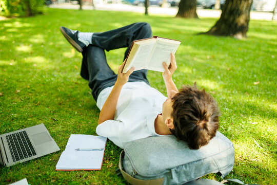 Relaxed teenager student reading a book while lying on the park grass.
