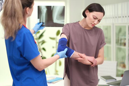 Female patient holds painful arm while nurse in blue scrubs checks injury and wraps support bandage, with nurse, patient, injury, arm detail, showing bandage, clinic, medical, healthcare context