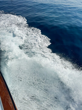 Close-up of a white foamy wave splashing and churning against dark blue sea water, created by a boat's wake, with fine sea spray droplets visible.