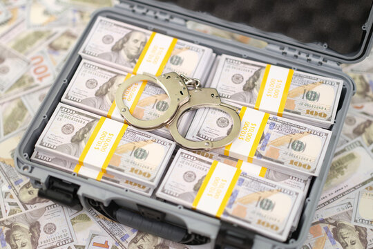 Open hard case packed with bundled US dollar bills, with metal handcuffs placed on top, creating strong visual themes of arrest, seized money, financial crime, corruption, and law enforcement