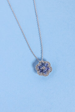 Elegant silver-tone necklace with a flower-shaped pendant featuring blue gemstone-style petals and sparkling clear accents, photographed on a pastel blue background.