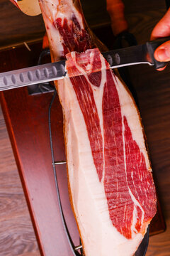 Traditional cured ham leg being sliced by hand with a long carving knife, close-up view of artisan charcuterie preparation.