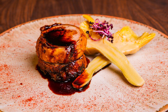 Elegant restaurant dish with glazed roasted meat medallions, braised vegetables, dark reduction sauce and fresh microgreens on a ceramic plate.