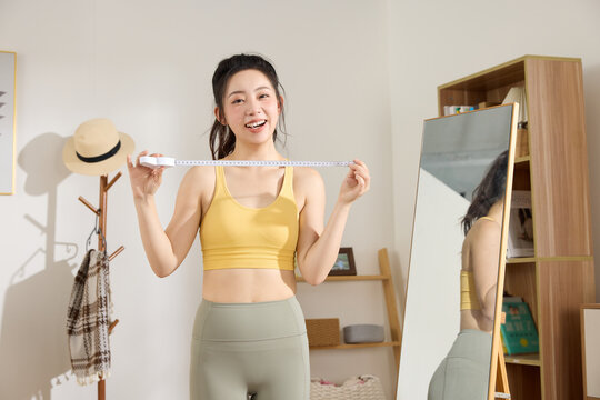 Happy woman measuring waist with tape measure in sportswear at home