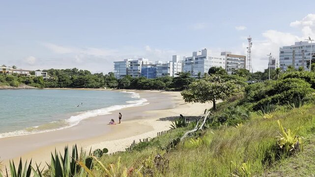 View of a sandy beach with turquoise water, modern buildings, and lush coastal vegetation in Guarapari, Espirito Santo, Brazil.