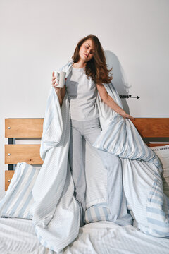 Young woman in gray pajama sits on a bed, wrapped in a striped blanket, holding a mug with a relaxed morning vibe. Studio vibe, cozy morning.