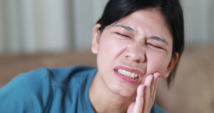 Woman suffering from toothache, holding her cheek in pain, tooth pain, dental problems, oral health, tooth sensitivity, cavity, dental treatment, and healthcare, close-up, Asian Thai people