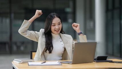 Fototapeta premium Excited woman cheering with fists raised, achieving business success while working on a laptop in a modern office