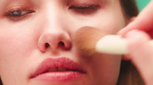 Makeup artist applying highlighter with brush to young teenager student face, beauty close up on pink background, cosmetics advertising for youth