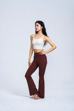 Young East Asian woman in white sports top and brown high-waisted yoga flare pants posing in plain light grey studio 