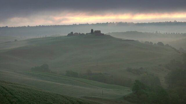View of rolling hills of Tuscany covered in morning mist with farm buildings on the horizon under a soft sunrise sky in Tuscany, Italy.