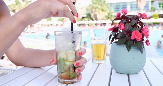 A sunny day unfolds as someone stirs a refreshing drink at a poolside table adorned with cheerful flowers. A second drink sits nearby, completing the relaxing atmosphere.