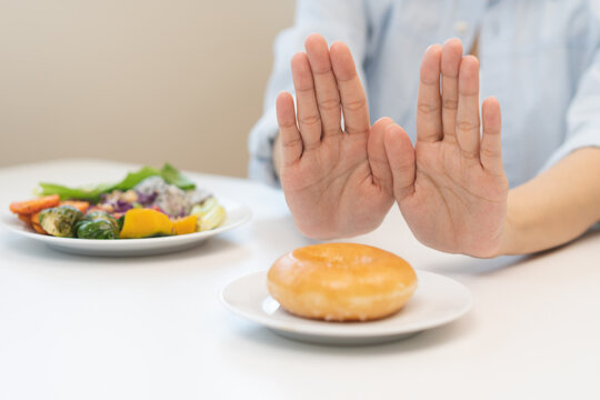 Diet concept, close up young woman, girl using hand push out, stop sweet donut, dessert or junk food on plate, choose green vegetables salad, eat low fat for good health. Female getting weight loss.