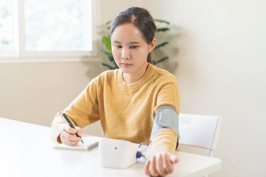 Asian young woman, girl writing blood pressure test results in a notebook while measuring with a digital monitor cuff at home, healthcare tracking and hypertension management concept.