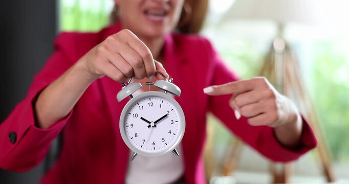 A woman in a stylish red blazer engages with an alarm clock, illustrating the significance of time while sharing a moment of reflection in a bright, cozy setting.