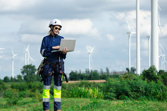Female wind turbine technician uses a laptop at a wind farm, wearing safety equipment while monitoring renewable electricity infrastructure and sustainable clean energy power systems outdoors.