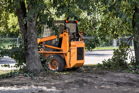 A small orange excavator or loader is working near trees in a suburban area. It is clearing brush and debris from the ground. Compact landscaping equipment
