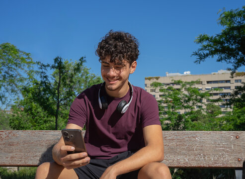 A young man sitting on a wooden bench outdoors on a sunny day, wearing wireless headphones and staring intently at his smartphone screen while smiling relaxed
