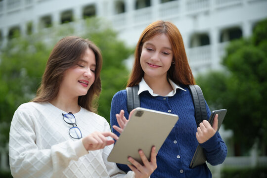 Two female students using a digital tablet together outdoors on campus, representing university education, online learning, technology and academic life.