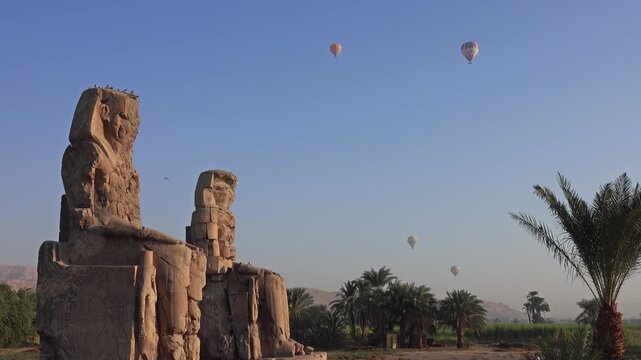View of the ancient Colossi of Memnon statues with hot air balloons floating in the sky over the landscape of Luxor Governorate, Egypt.