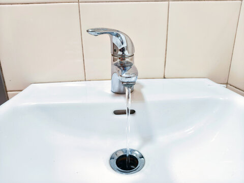 Chrome faucet with running water flowing into white ceramic washbasin. Bathroom sink with drain hole and overflow slot. Plumbing and water supply. Hygiene and sanitation concept