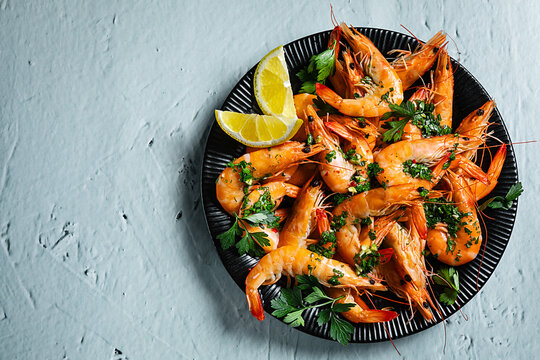 Fresh cooked shrimp with garlic, parsley and lemon on a black plate, top view, dark background.