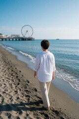 Fototapeta premium Mature woman in white linen shirt walks on sandy beach. Back view of lady strolling along ocean shore near seaside pier. Large ferris wheel stands on pier under blue sky. Peaceful summer vacation,