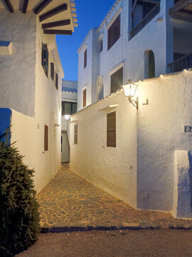 Estrecho callej&oacute;n empedrado de noche, flanqueado por casas tradicionales de fachadas blancas encaladas, iluminadas por un farol c&aacute;lido bajo un cielo de un azul profundo de crep&uacute;sculo.
