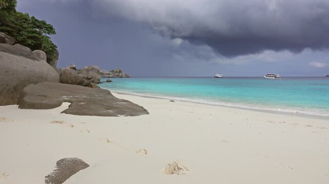 View of Similan Islands beach with white sand, large granite boulders, turquoise water, and boats under a dark stormy sky in Lam Kaen, Phang Nga, Thailand.
