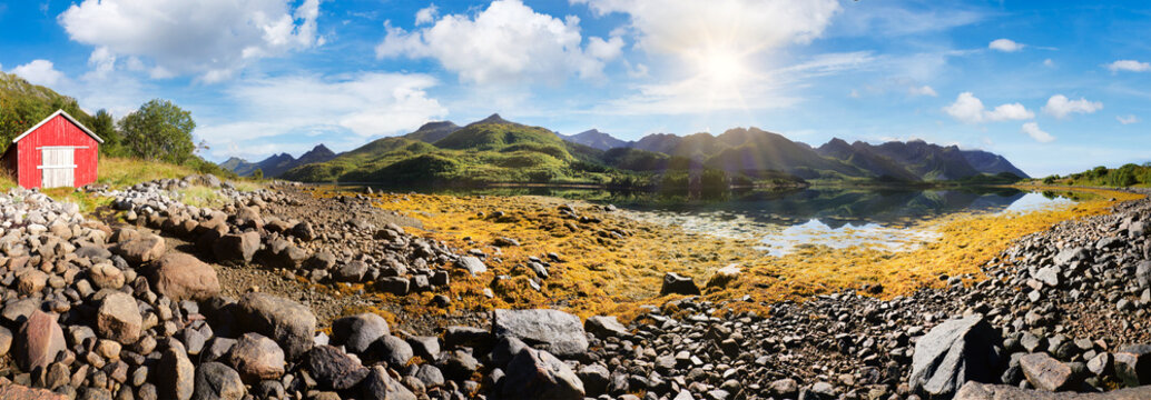 Picturesque coastal landscape near Olderfjord on the Lofoten Islands with rocky shore, calm water, green mountains and red wooden house in sunny weather under blue sky.