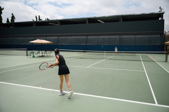 Rear view of Asian Woman Playing Outdoor Tennis Match on Green Hard Court with Opponent Across Net