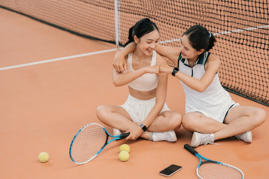 Two Asian Women Friends Taking a Break on Tennis Court, Chatting and Fist Bumping Together