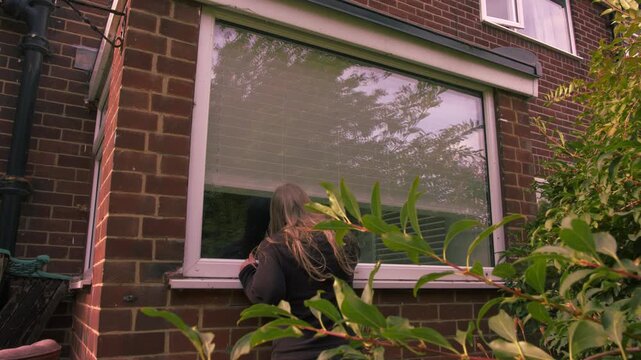 Woman looking through house window with blinds through view of trees