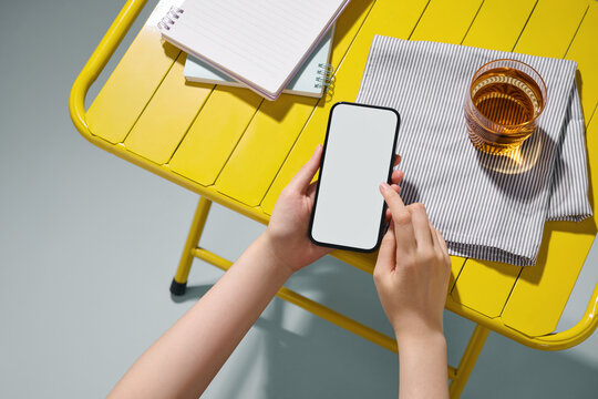Hands holding a smartphone with a blank screen on a yellow table.