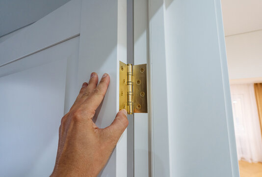 Closeup hand inspecting brass door hinge on white interior door, fingers touching hinge screws and door jamb seam during home maintenance in bright natural light detail