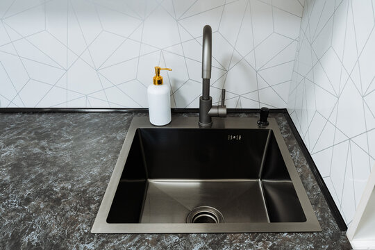 Overhead view of modern matte black kitchen sink with tall curved faucet and white pump dispenser on dark stone countertop with geometric tile backsplash