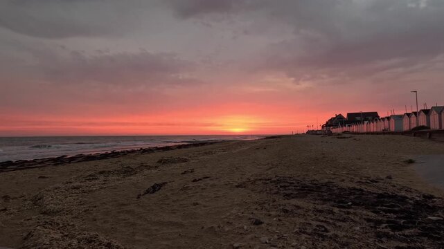 View of the vibrant sunset over the sandy beach with rows of beach huts along the coastline in Bernieres-sur-Mer, Normandy, France.