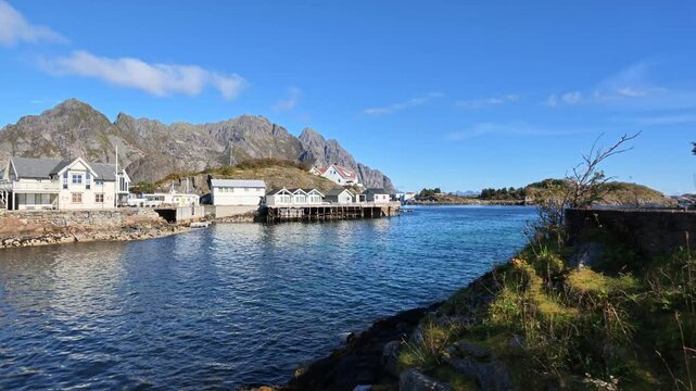 Traditional houses by the water in Henningsv&aelig;r on the Lofoten Islands with rocky mountains, blue sea and coastal vegetation under sunny blue autumn skies in Northern Norway