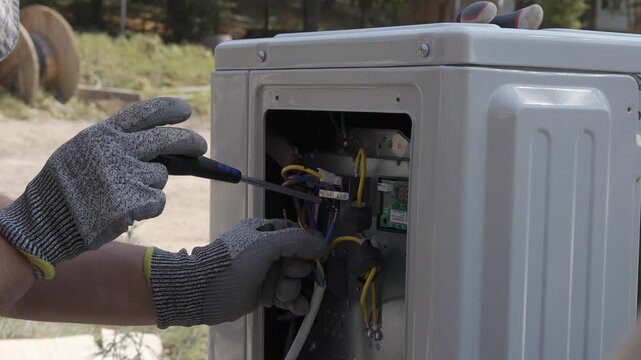 Close-up of an HVAC technician in protective gloves connecting electrical wires with a screwdriver inside an outdoor air conditioner unit. Professional split system installation and wiring process 