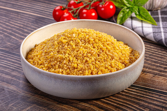 Bowl of golden bulgur grain on dark wood table with cherry tomatoes, basil, and checkered napkin in background