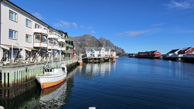 Picturesque harbor view of Henningsv&aelig;r on the Lofoten Islands with fishing boat, traditional wooden houses, calm blue water and impressive rocky mountains under sunny autumn sky.