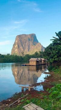 A large, flat-topped mountain view stands out in Muang Fuang, Laos. There is accommodation on a wooden raft in the middle of the water, surrounded by lush greenery on a clear day.