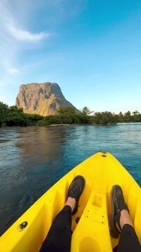 A video shot from a yellow kayak, showing the occupant's feet. The kayak is floating on turbulent water against a backdrop of flat-topped mountains and forest in Muang Fueng, Laos. 