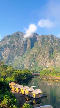 An aerial panning shot reveals a long mountain range surrounded by forest in Muang Fuang, Laos. There is accommodation on a wooden raft in the middle of the water on a clear day with a blue sky.
