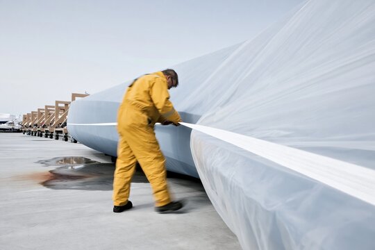 Shrink-wrapped boat hull being secured by a man in yellow oilskins on a wet boatyard for marine insurance and storm preparedness