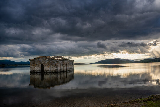 View of the submerged St. John of Rila church standing in the calm waters of the Zhrebchevo Reservoir under a dramatic stormy sky with sunbeams breaking through in Sliven, Bulgaria.