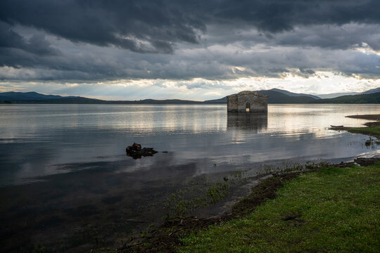 View of the submerged Church of St. John of Rila in the Zhrebchevo Reservoir under a dramatic, dark, cloudy sky with soft light reflecting on the water surface in Sliven, Bulgaria.